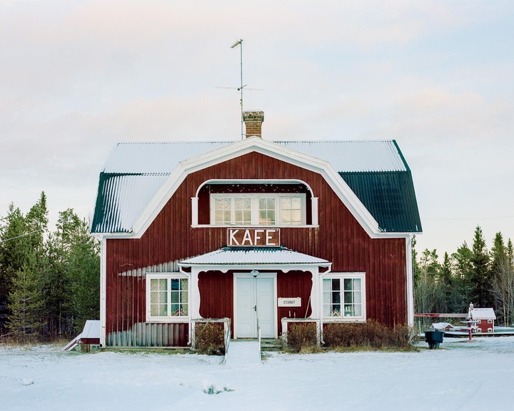 A red cafe building with a snow-covered roof stands in a snowy landscape surrounded by pine trees.