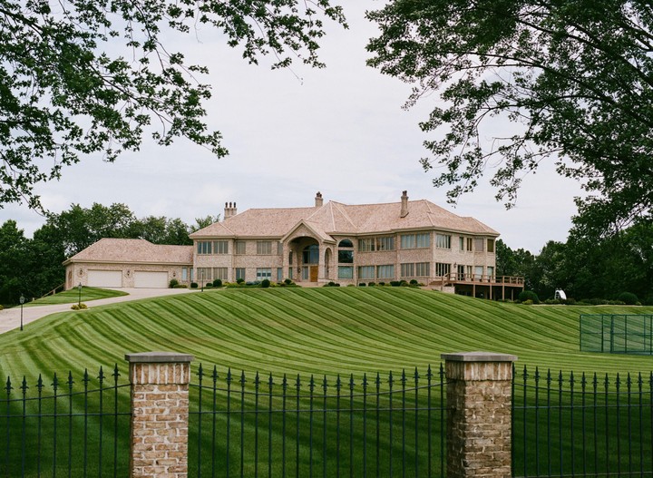 A large house with a manicured lawn is seen through a wrought iron fence.