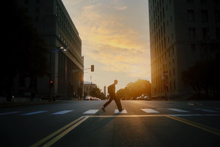 A man with a skateboard crosses a street at sunset between two tall buildings.