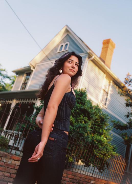 A woman stands in front of a house with a brick wall and metal fence in the foreground.