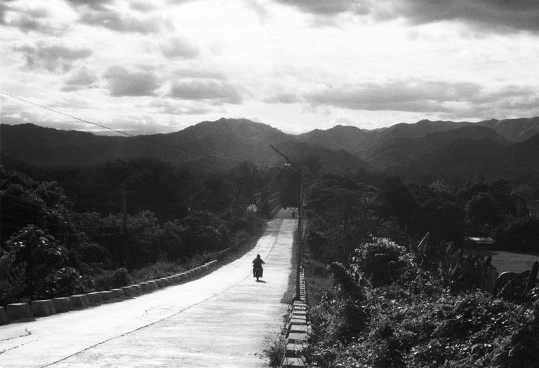 A person rides a motorcycle down a long road surrounded by mountains and trees under a cloudy sky.
