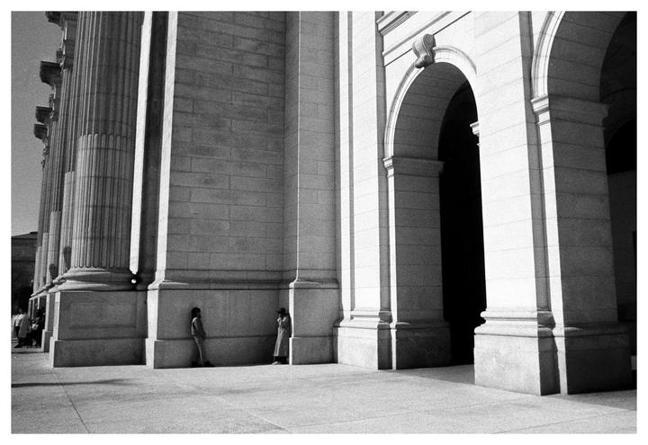 Two people stand in the shadow of a large stone building with columns and arches.