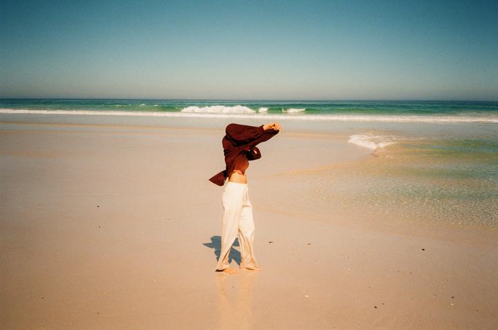 A woman stands on a sandy beach with her shirt pulled over her head, facing the ocean.