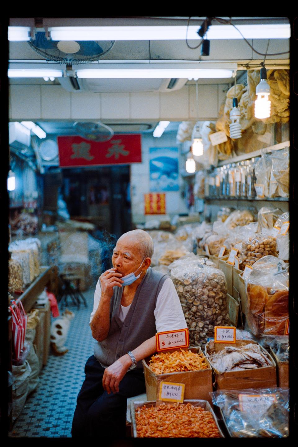 A man with a face mask under his chin smokes a cigarette while sitting in a market stall surrounded by dried goods.