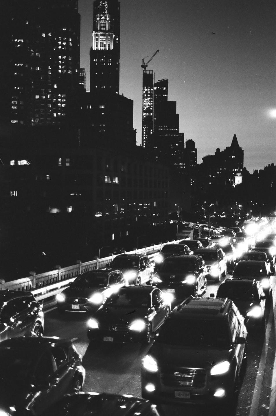 A long line of cars with their headlights on are stuck in traffic on a bridge at night, with city buildings in the background.