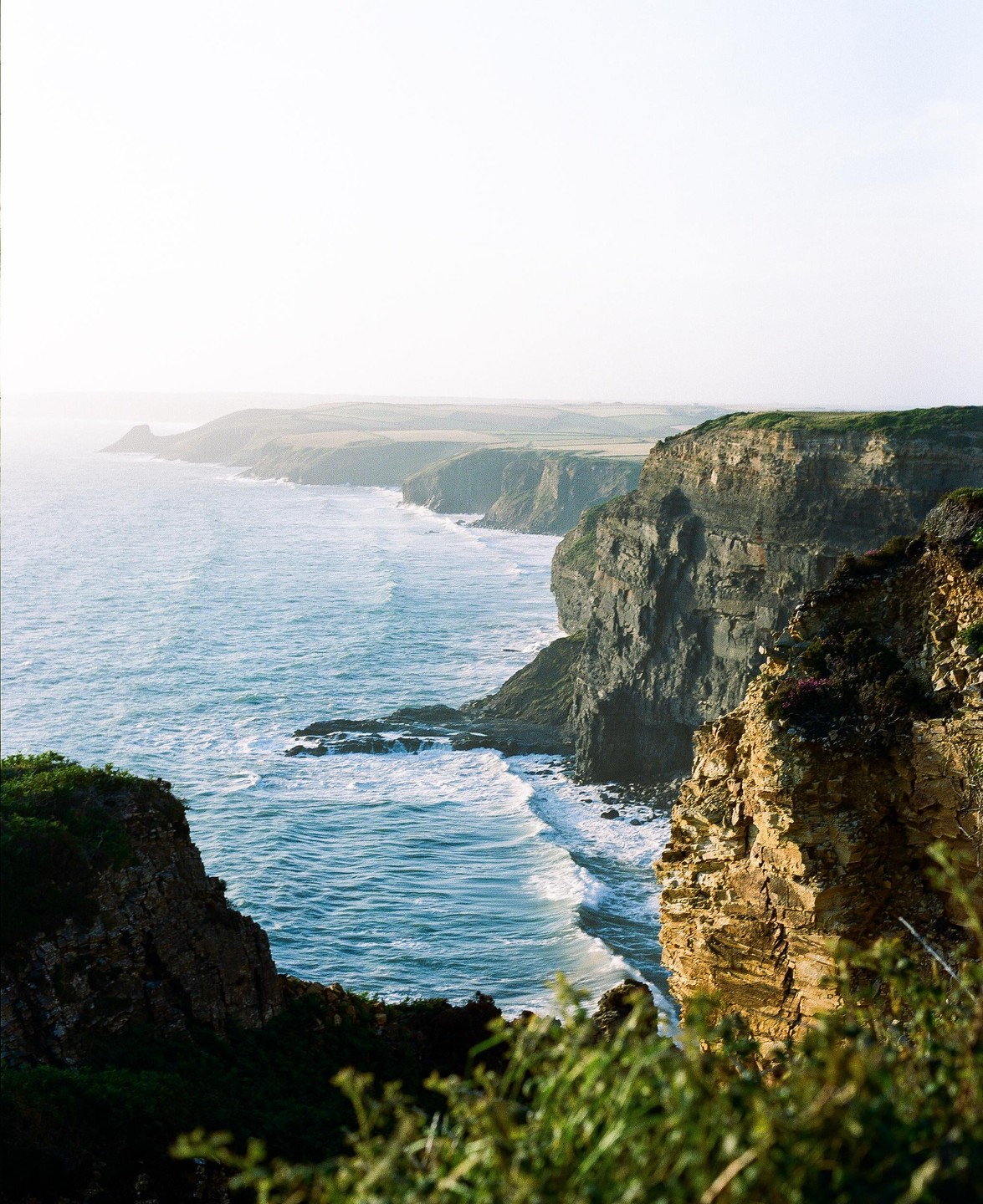Waves crash against a rugged coastline with cliffs and distant fields under a bright sky.