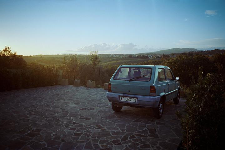 A teal car is parked on a stone patio overlooking a valley with vineyards and a town in the distance at sunset.