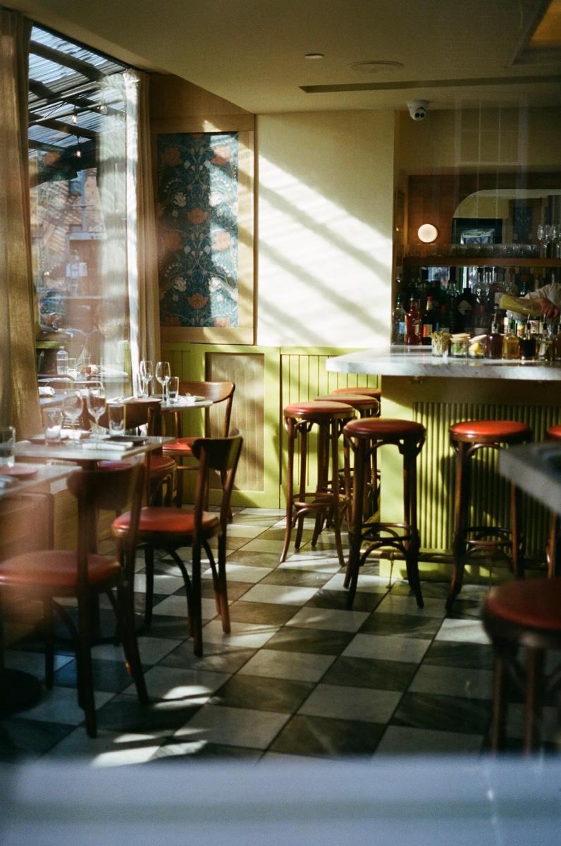 Sunlight streams into a restaurant with checkered floors, tables, and a bar with stools.