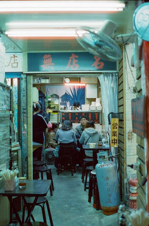 People sit at small tables in a narrow restaurant with a neon sign and a ceiling fan.