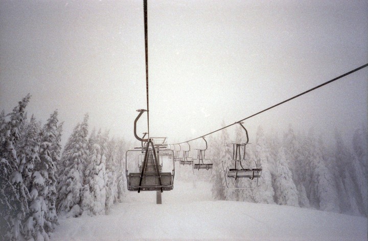 Empty ski lift chairs ascend a snowy mountain with snow-covered pine trees on either side under a hazy sky.
