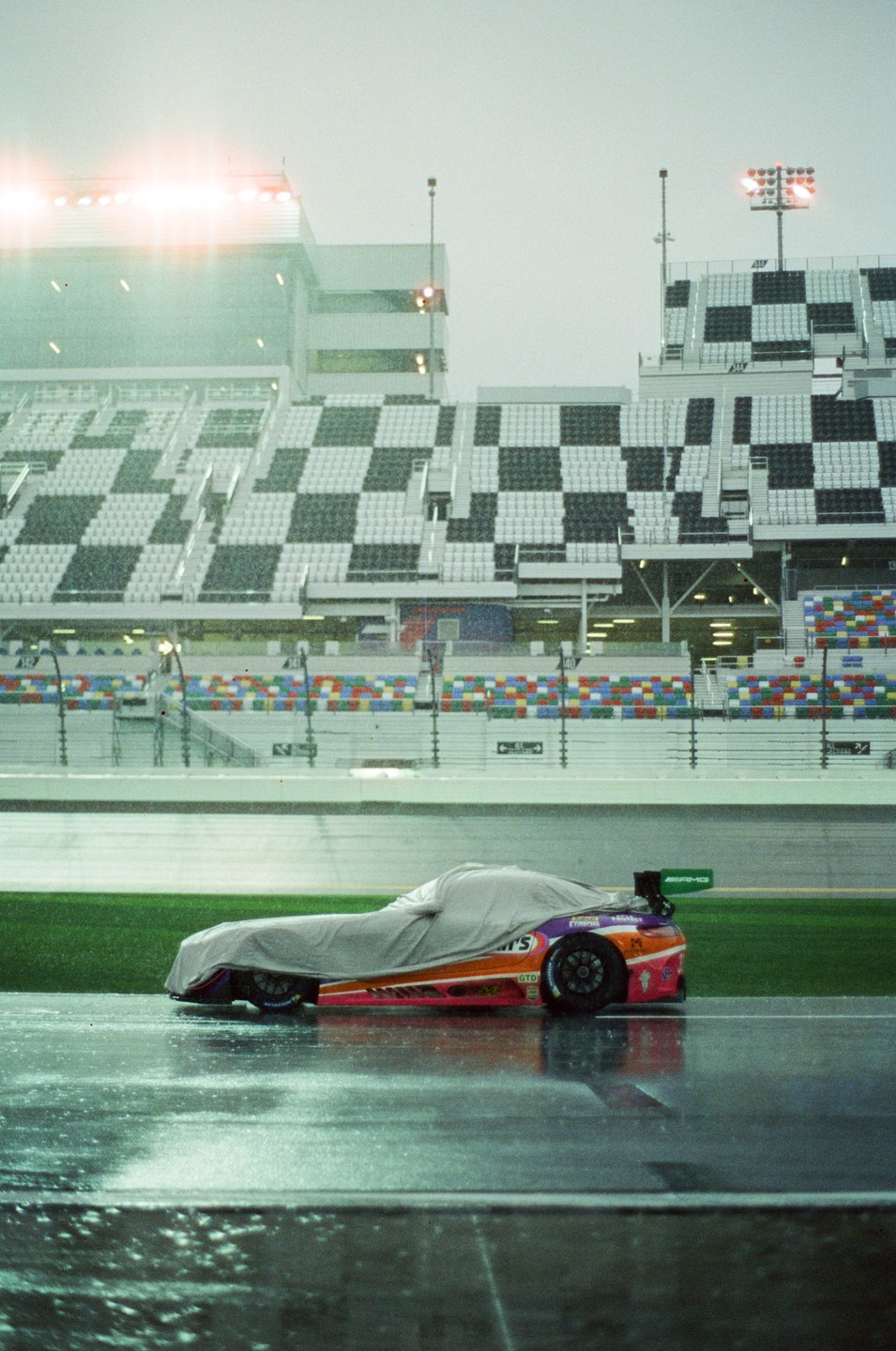 A partially covered race car sits on a wet track in front of stadium seating.