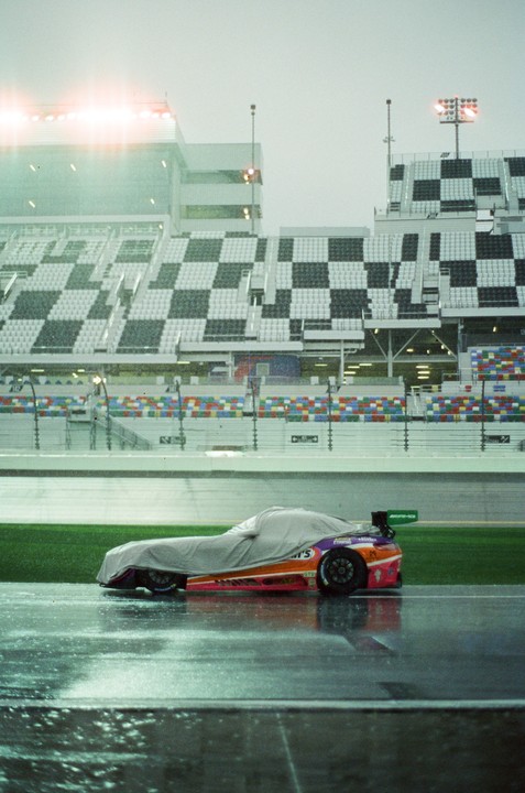 A partially covered race car sits on a wet track in front of stadium seating.