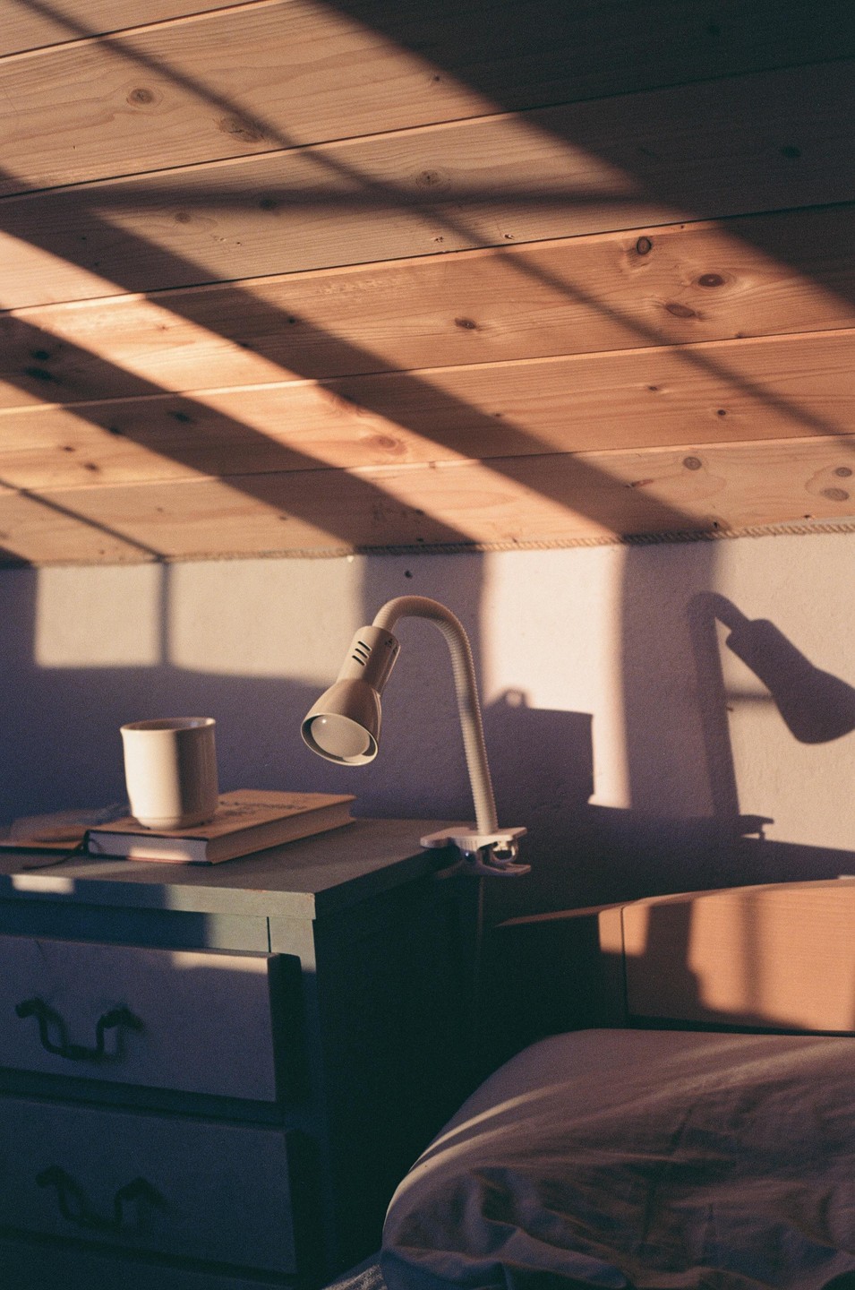 Sunlight streams through a window, casting shadows on a wooden ceiling and a bedside table with a lamp, mug, and book.