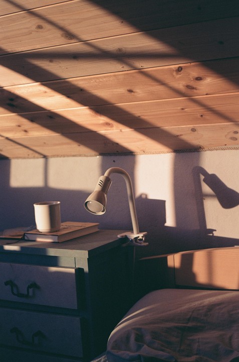 Sunlight streams through a window, casting shadows on a wooden ceiling and a bedside table with a lamp, mug, and book.