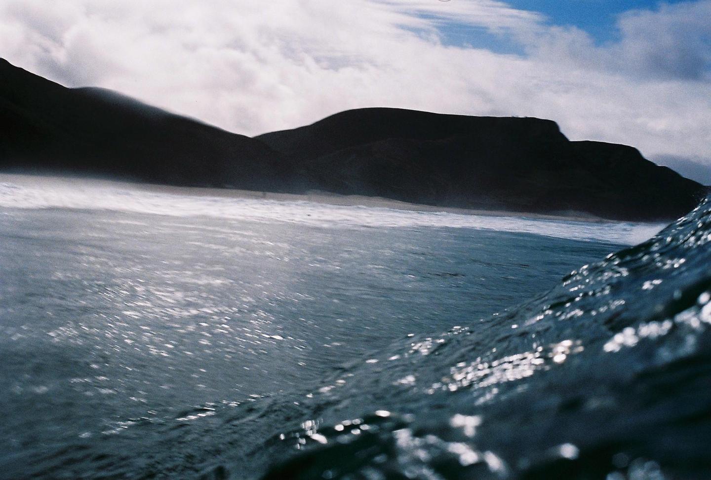 A low-angle view from the ocean shows waves breaking on a beach with dark mountains in the background under a cloudy sky.