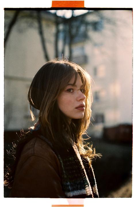 A woman with long hair and a scarf looks to the side with a building in the background.