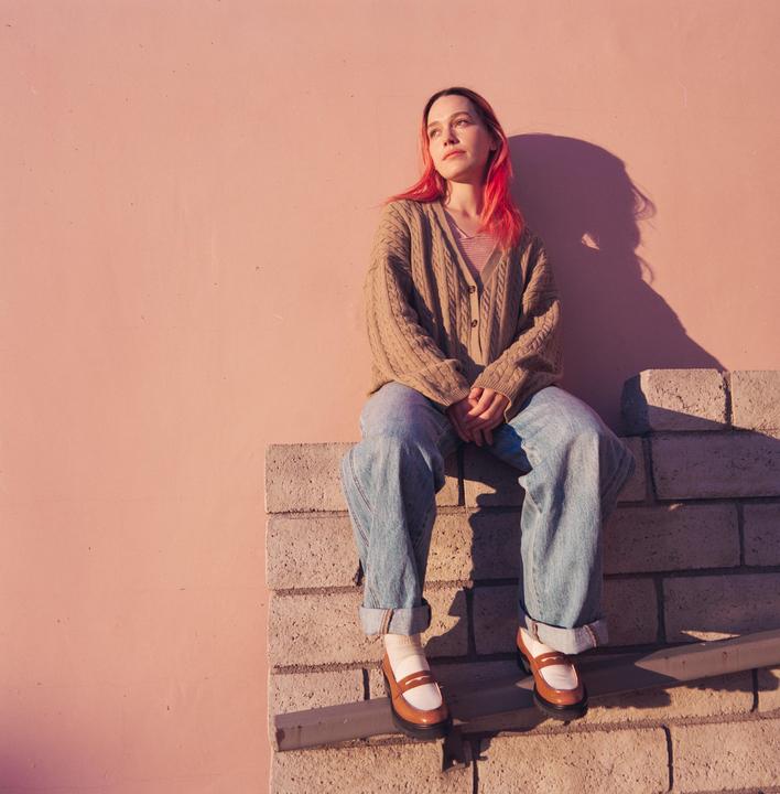 A woman with pink hair sits on a brick wall in front of a pink wall.