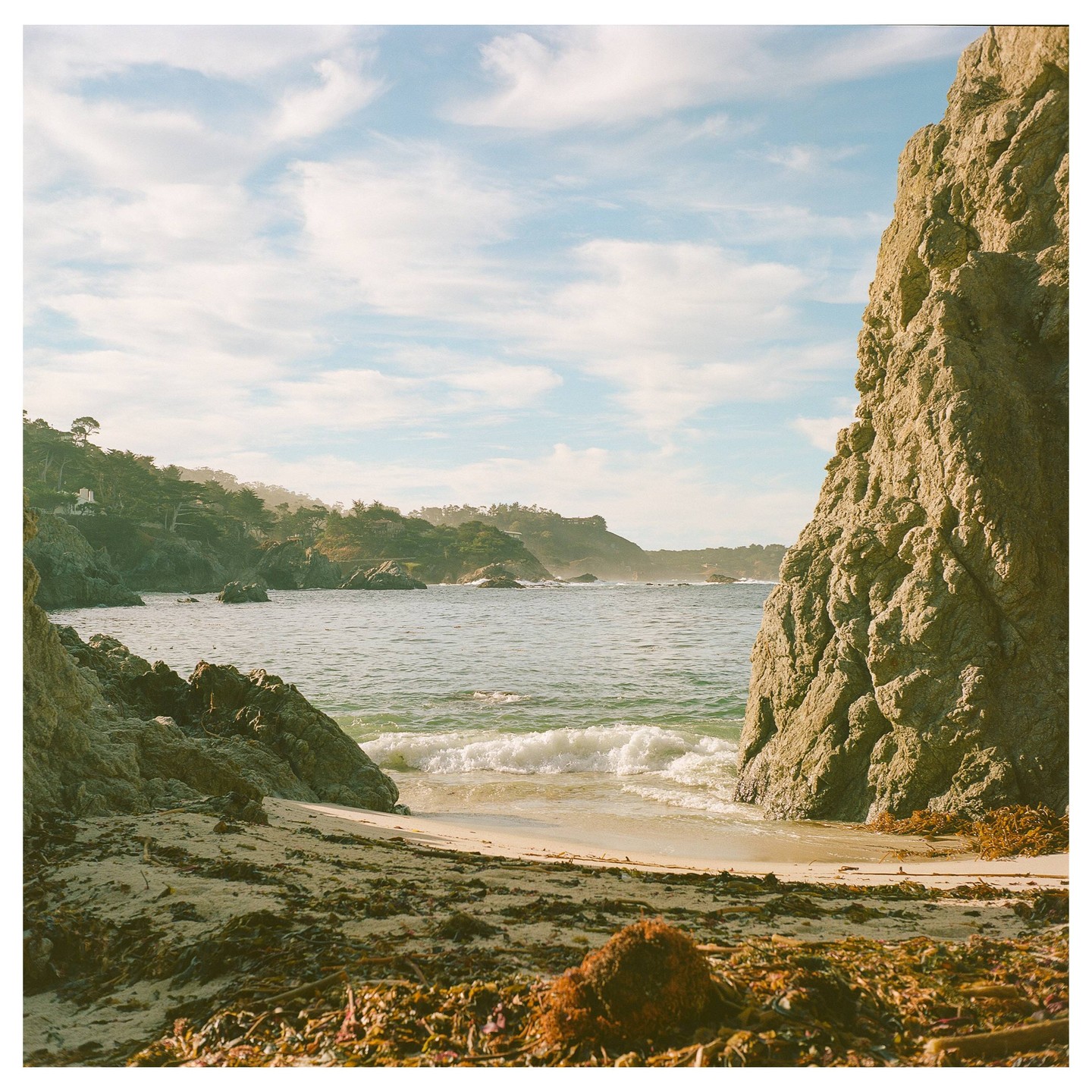A small sandy beach is visible between two large rock formations with the ocean and a distant tree-covered coastline under a cloudy sky.