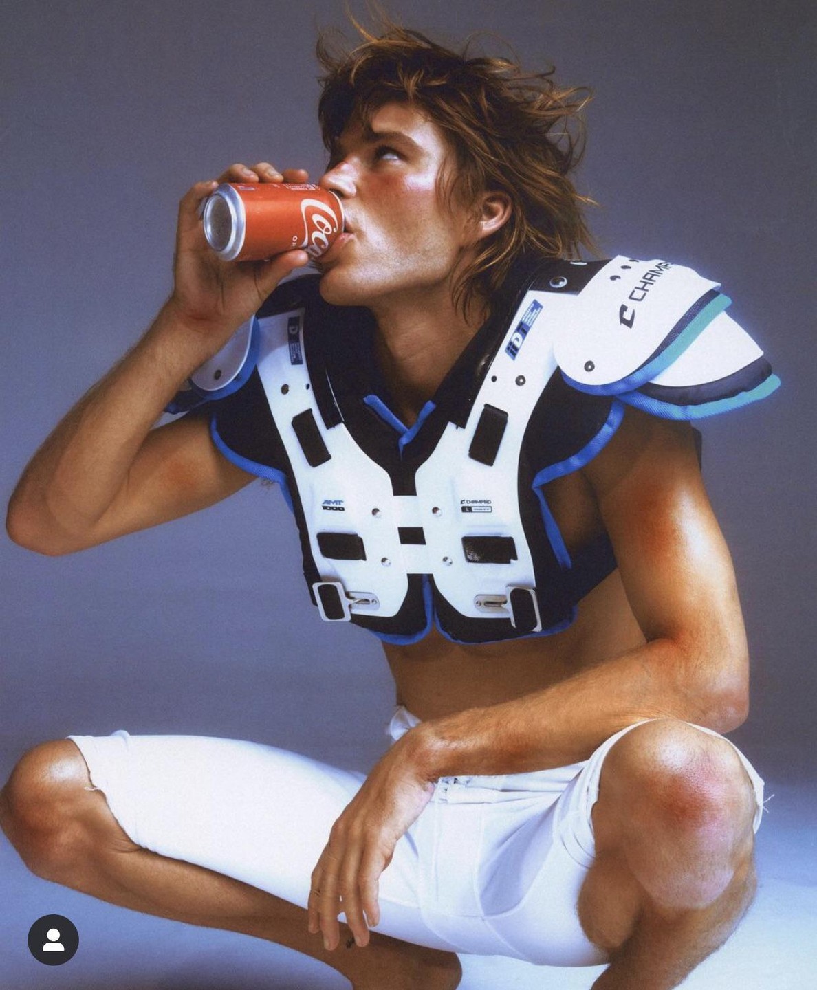 A man in football pads and white pants crouches while drinking from a Coca-Cola can.