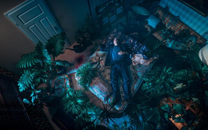 A man in a suit lies on a mattress surrounded by many plants in a dimly lit room.