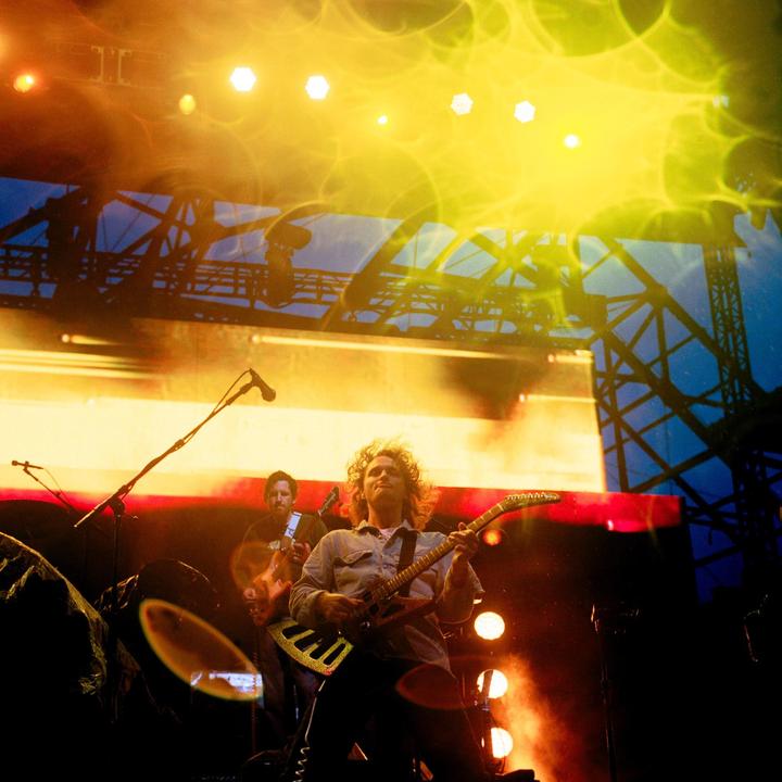 A man with curly hair plays an electric guitar on a stage with another man playing guitar in the background, illuminated by bright yellow and orange lights.