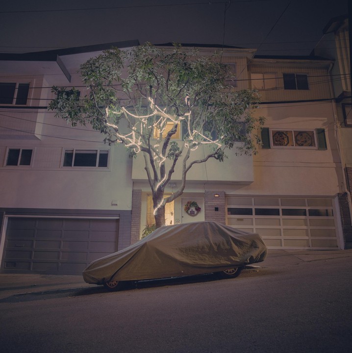 A car covered with a tarp is parked on a street in front of a house with a tree decorated with string lights.