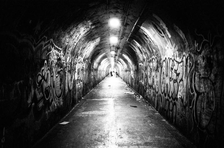 A long, dark tunnel with graffiti-covered walls leads to a bright light in the distance, where two figures are visible.
