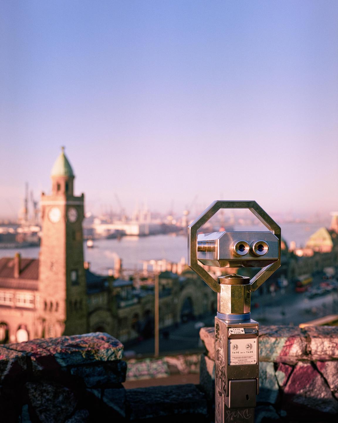 A coin-operated binocular viewer overlooks a harbor with a clock tower and ships under a clear sky.
