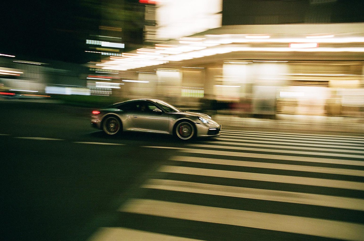 A silver car speeds across a crosswalk at night, with blurred lights in the background.