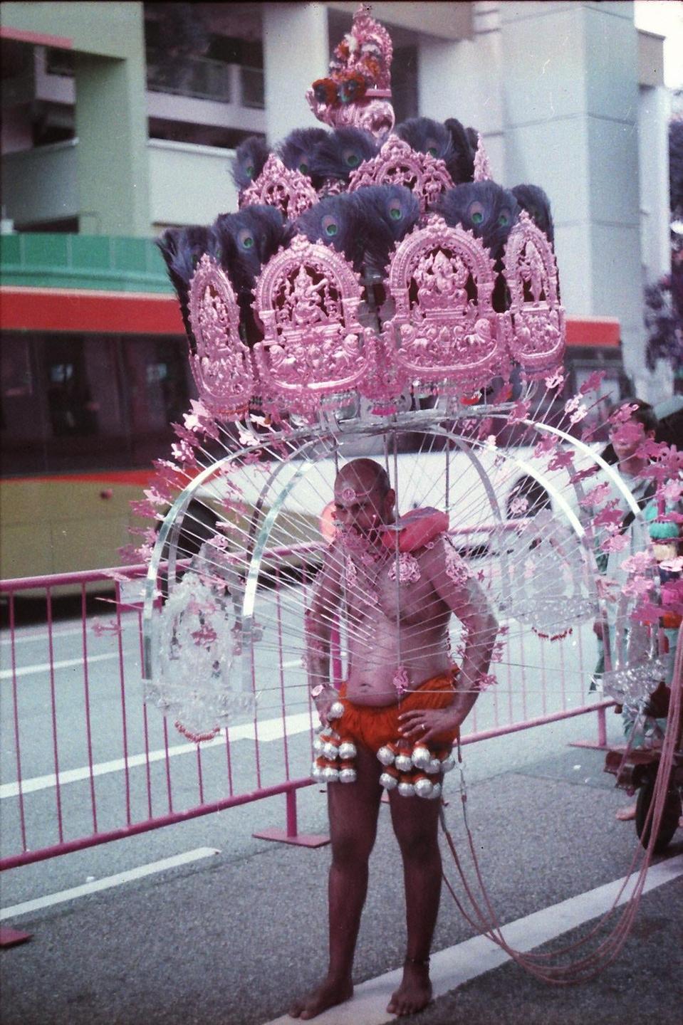 A man stands in the street wearing an elaborate, decorated kavadi with peacock feathers and pink ornaments.