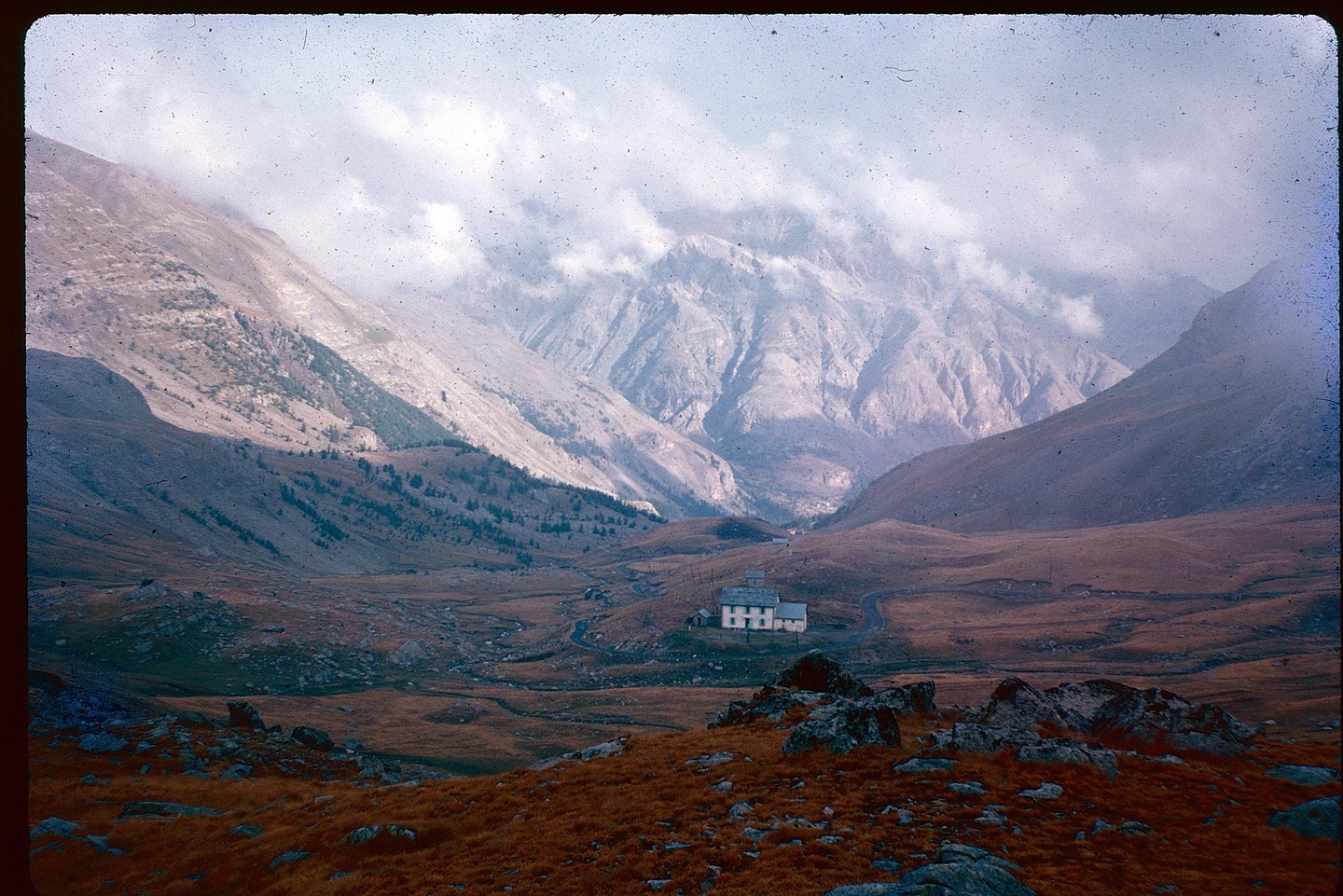 A small building sits in a valley surrounded by mountains under a cloudy sky.