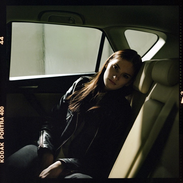 A woman with long hair and a leather jacket sits in the back seat of a car, looking towards the viewer.