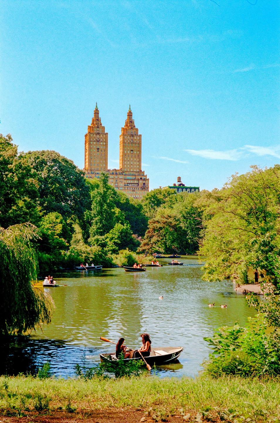 Two women row a boat on a lake with many other boats, surrounded by trees and buildings in the distance.