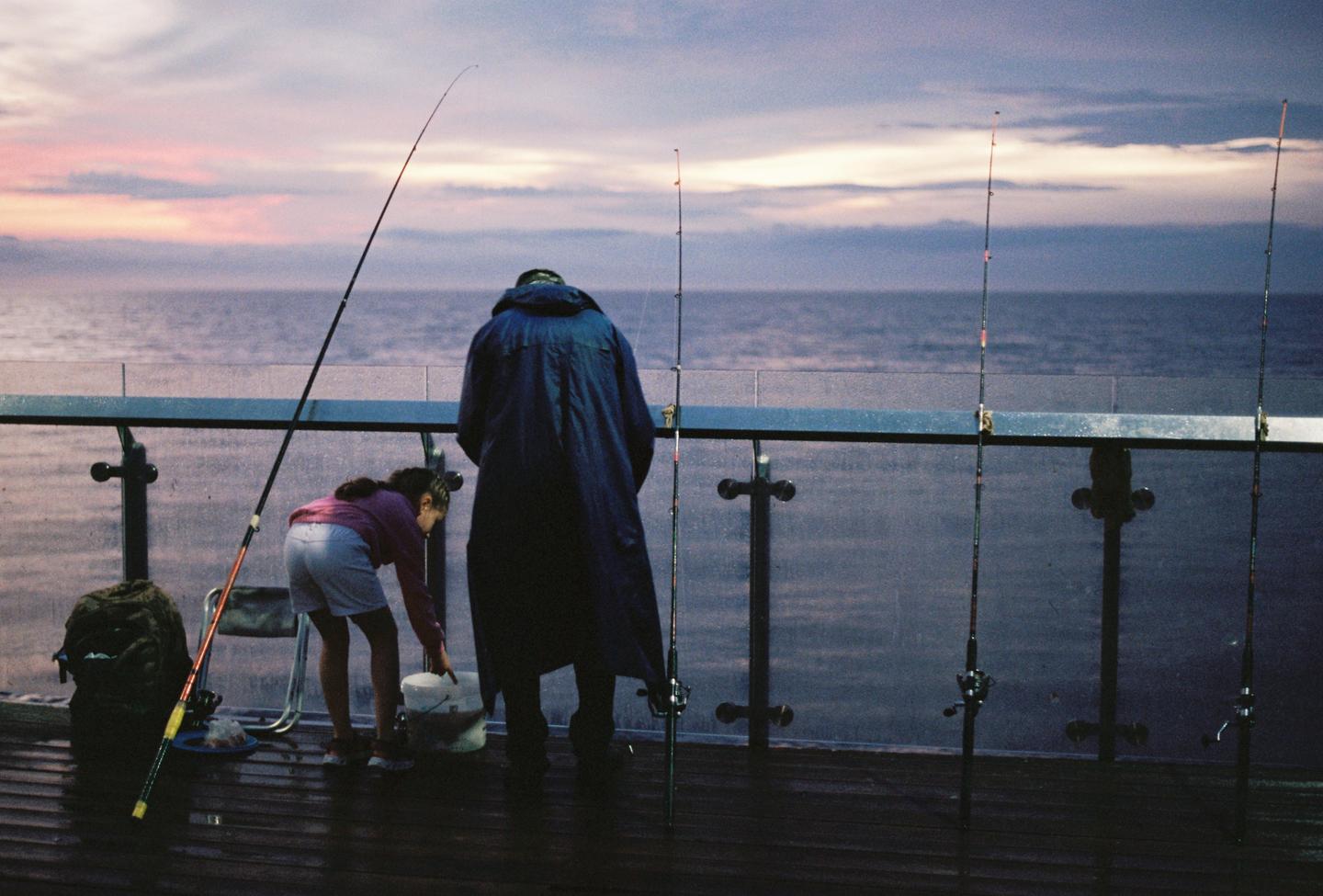 A man and a girl are fishing from a pier at dusk.