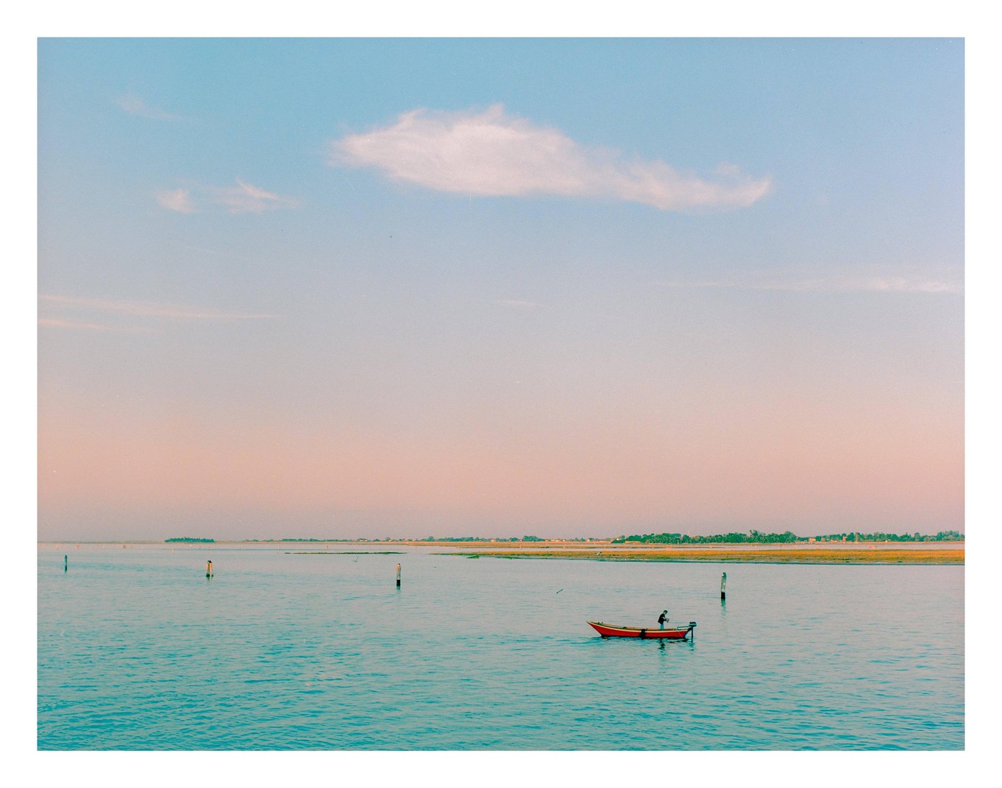 A person in a small red boat floats on the water with a distant shoreline under a pastel sky.