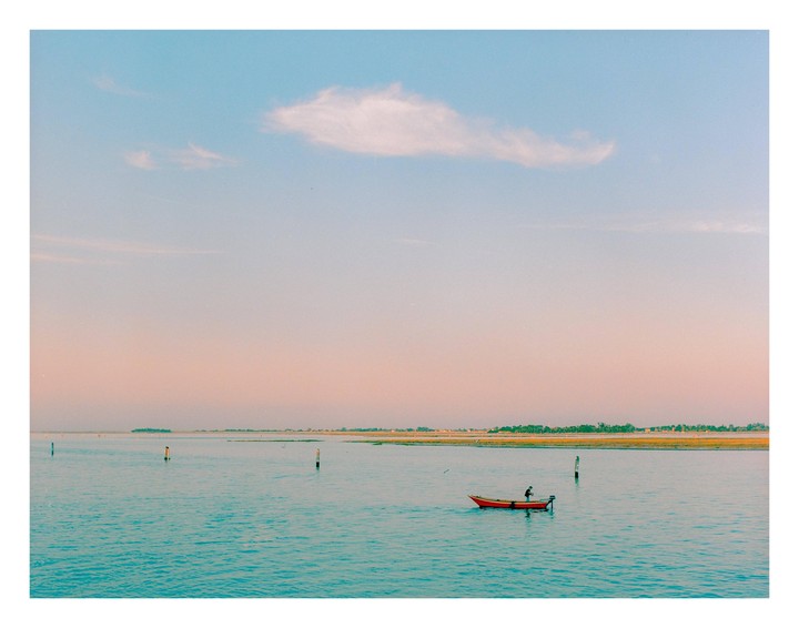 A person in a small red boat floats on the water with a distant shoreline under a pastel sky.