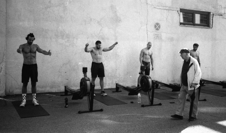 Four men are exercising outdoors with rowing machines next to a cracked wall with a window.