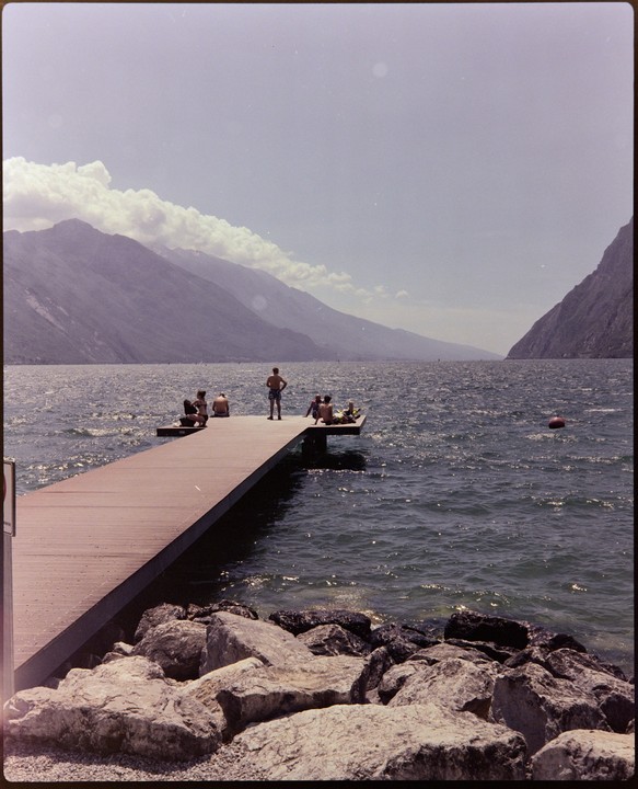 A group of people relax on a pier extending into a large lake surrounded by mountains.