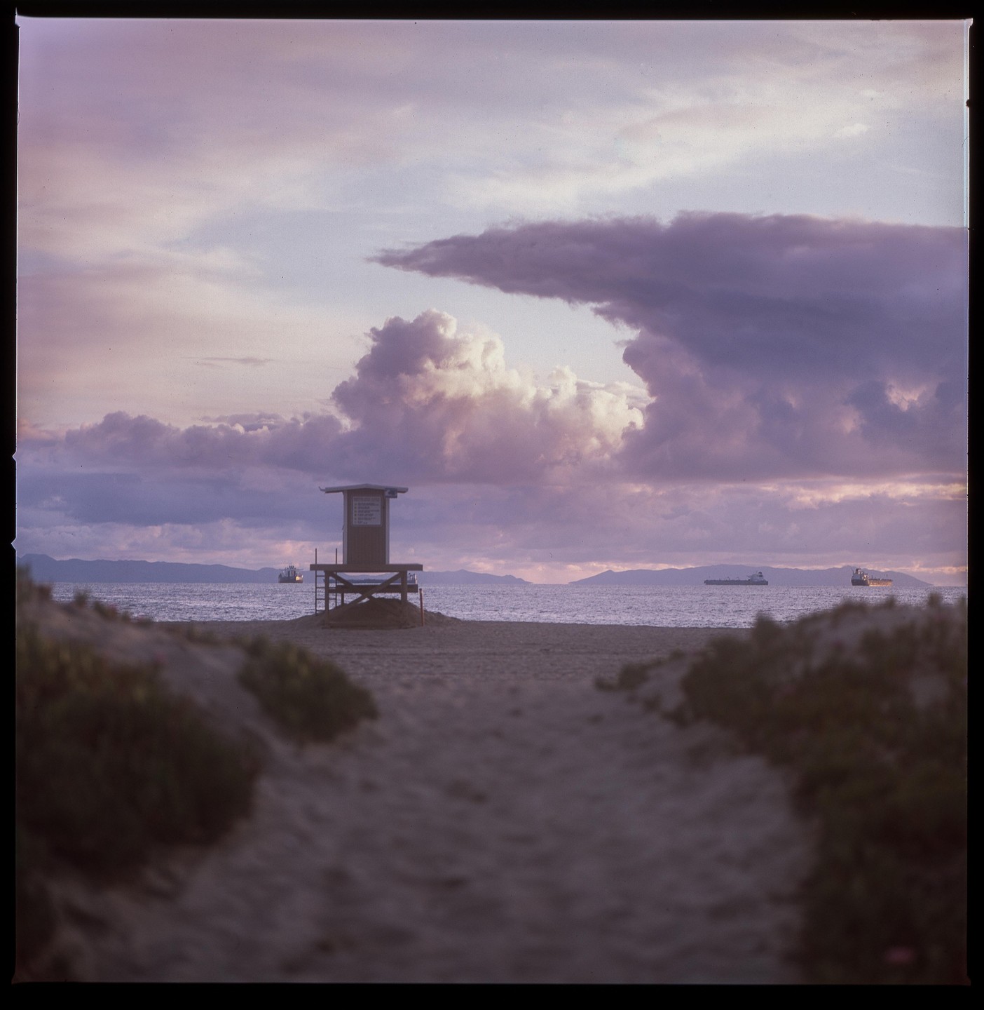 A lifeguard tower stands on a sandy beach under a dramatic, cloudy sky with ships visible on the horizon.