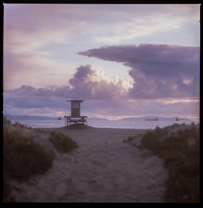 A lifeguard tower stands on a sandy beach under a dramatic, cloudy sky with ships visible on the horizon.