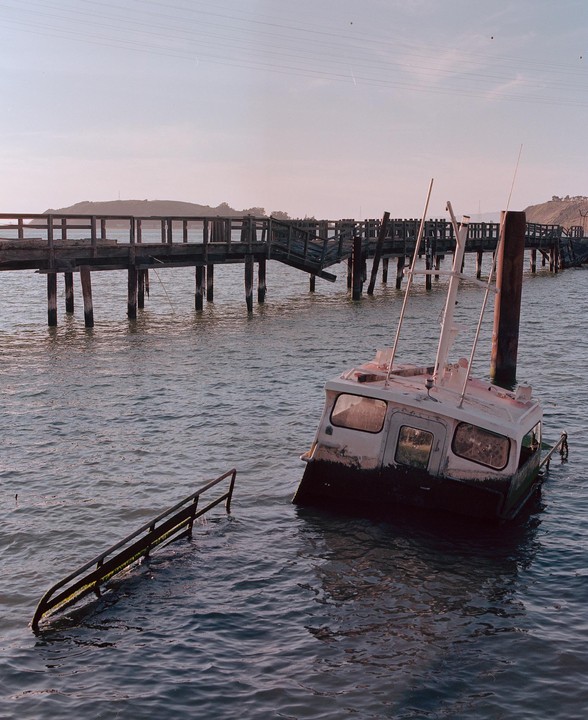 A partially submerged boat floats next to a broken wooden pier.