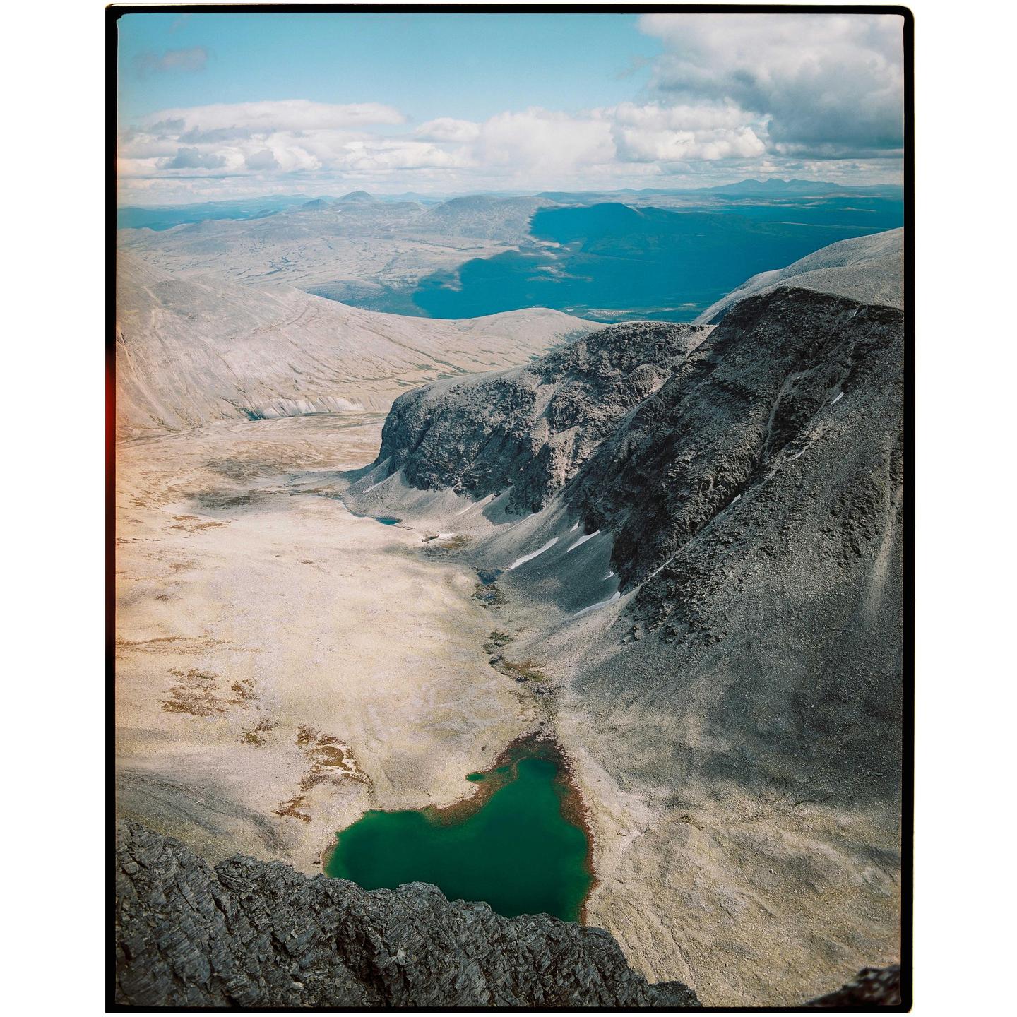 A high-angle landscape shows a valley with a bright green lake, surrounded by mountains and a distant body of water under a cloudy sky.