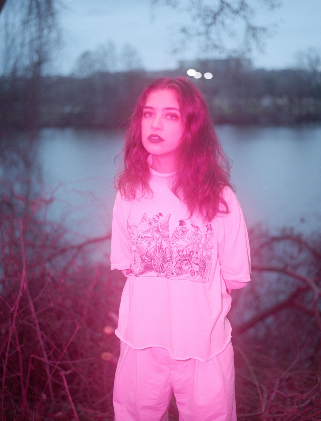 A woman with long hair and dark lipstick stands in front of a body of water, surrounded by pink-tinted foliage.