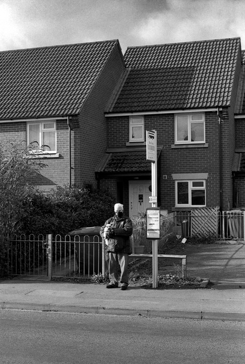 A man wearing a face covering holds a small dog while standing at a bus stop in front of brick houses.