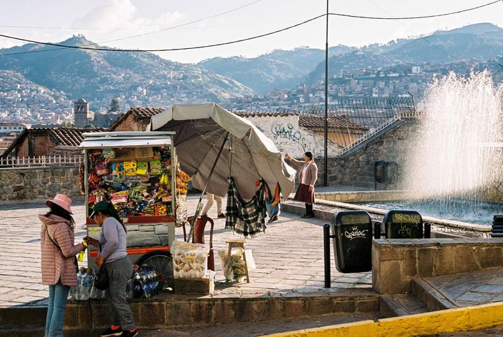 Two women stand next to a snack cart with a large umbrella and a fountain in the background, with a city on a mountain in the distance.