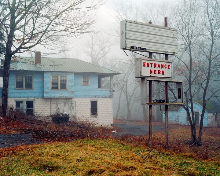 An abandoned motel with a blue and white building and a sign that says "Entrance Here" is shrouded in fog.