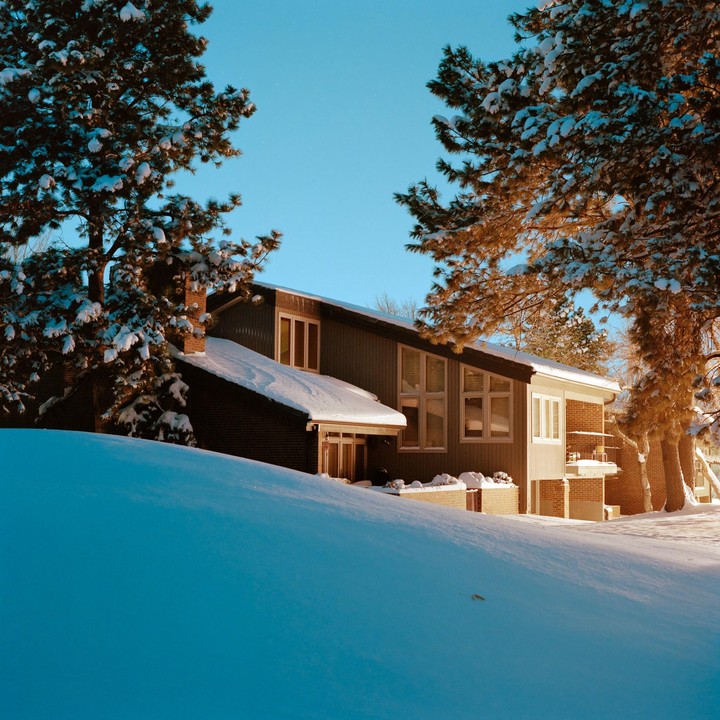 A house with snow-covered roofs and trees stands on a snowy hill under a clear blue sky.