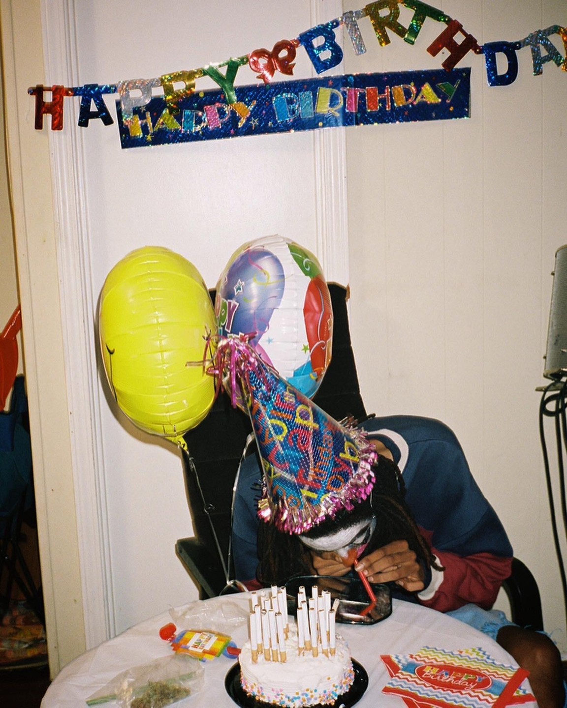 A person wearing a party hat and sunglasses is hunched over a table with a birthday cake, balloons, and a "Happy Birthday" banner in the background.