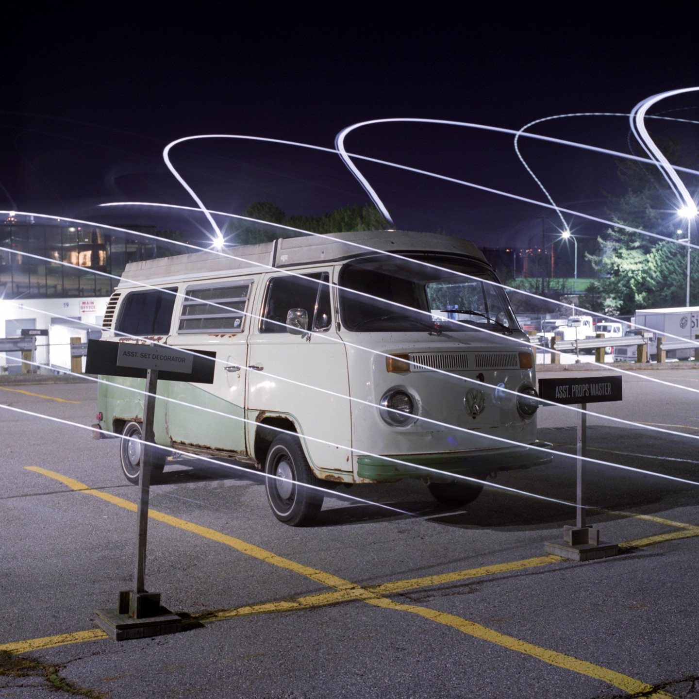 A light-painted van is parked in a lot at night, with two signs in the foreground.