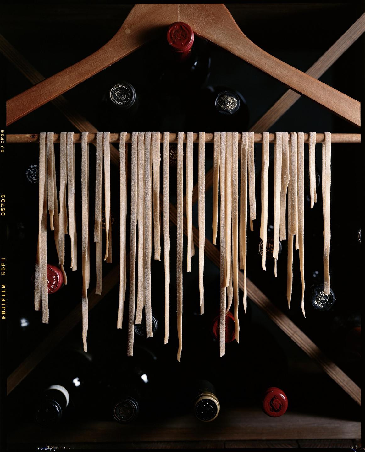 Fresh tagliatelle pasta hangs from a wooden hanger in front of a wine rack.